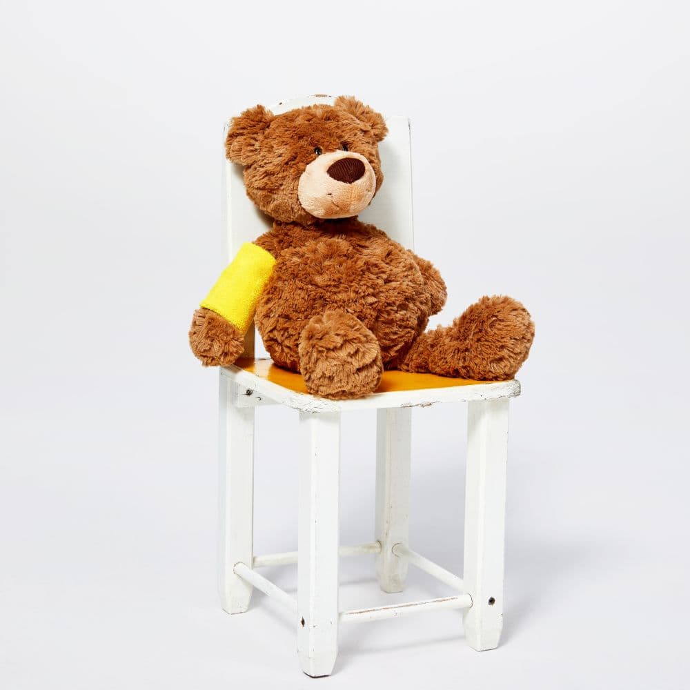 A brown teddy bear with a yellow arm bandage sitting on a white chair