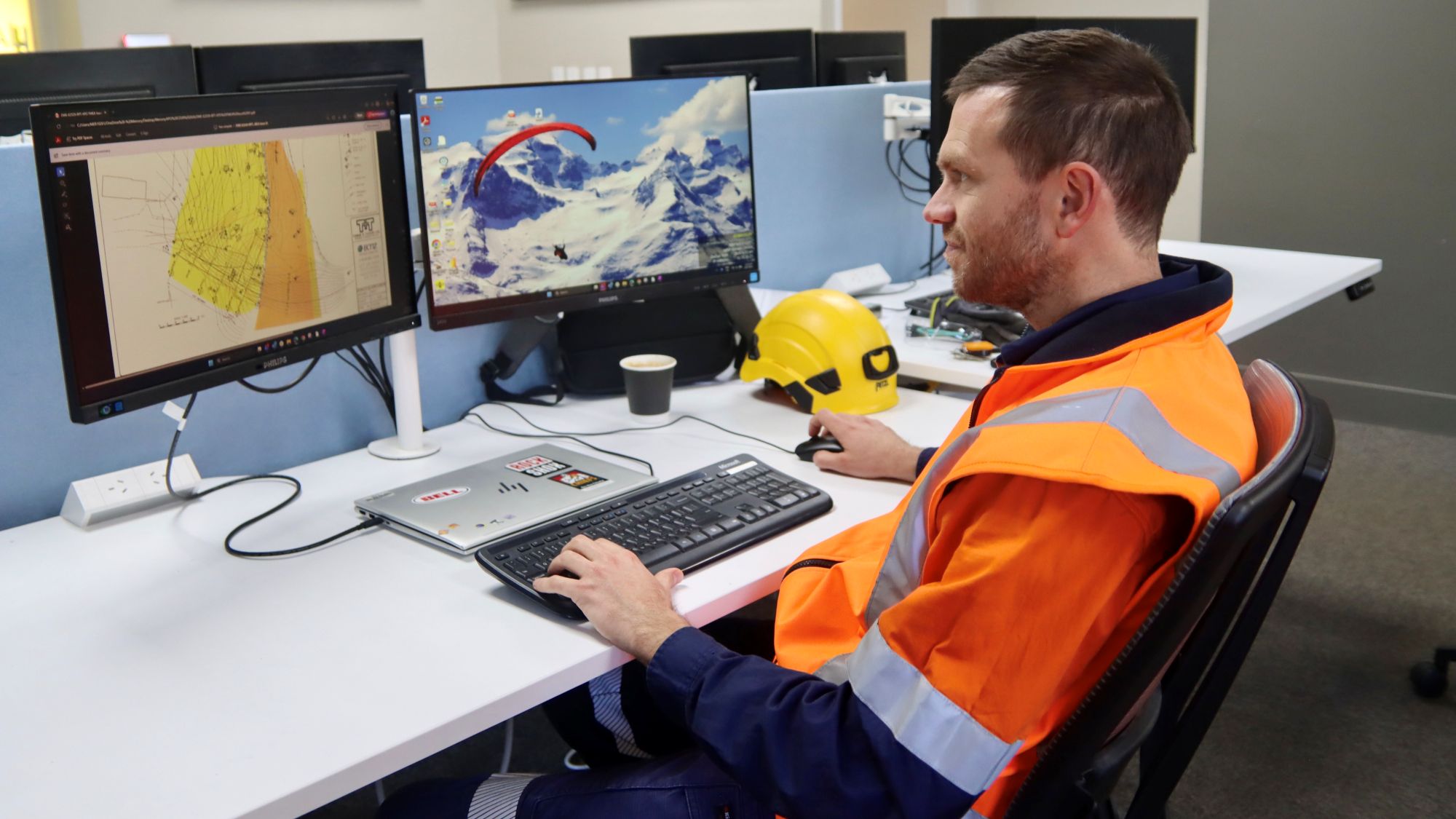 Man in orange hi-viz seated at desk operating a computer with two screens. 