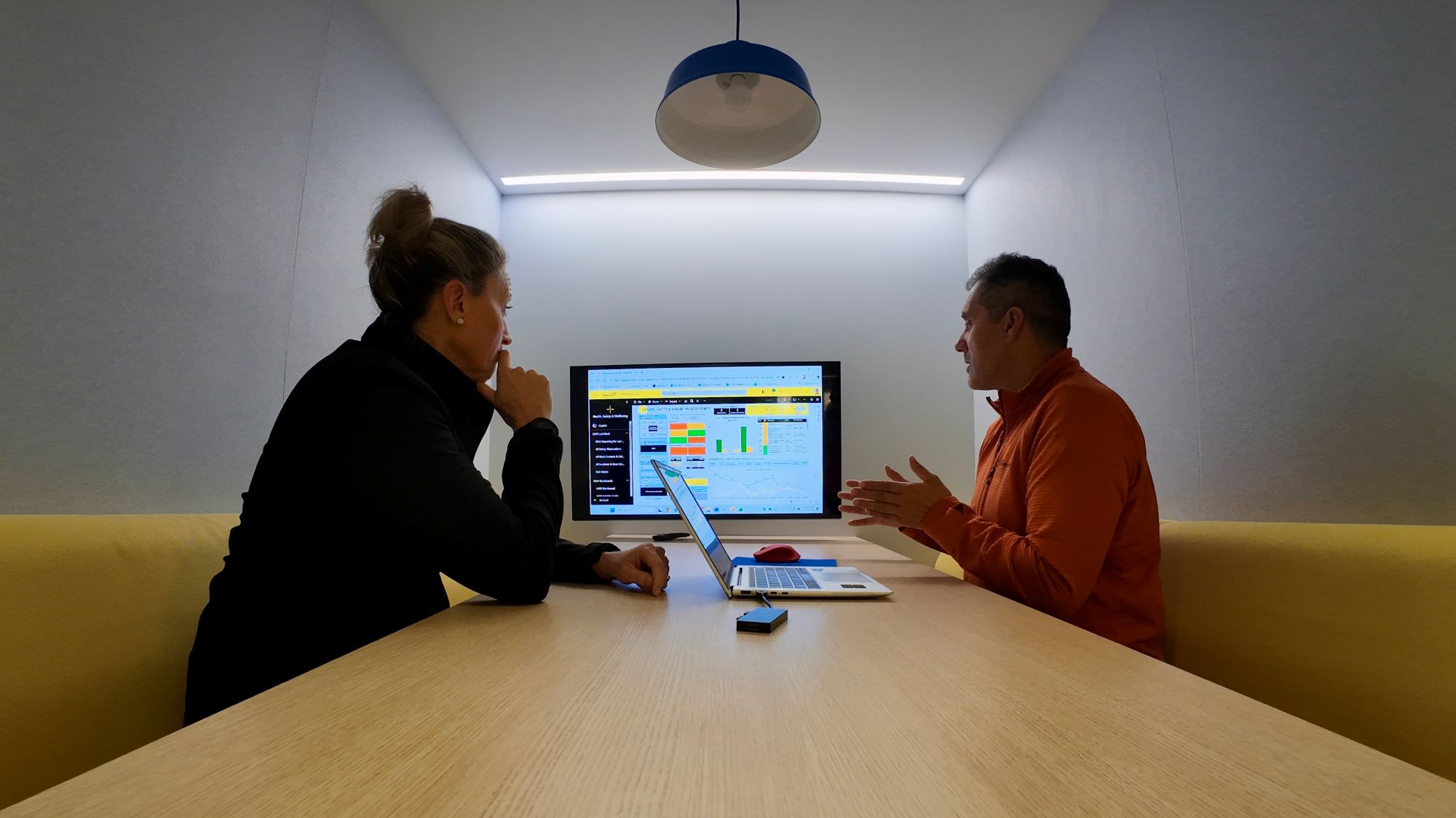 Two people sat in a collaboration booth in an office looking at a shared screen, with a laptop on the table between them.