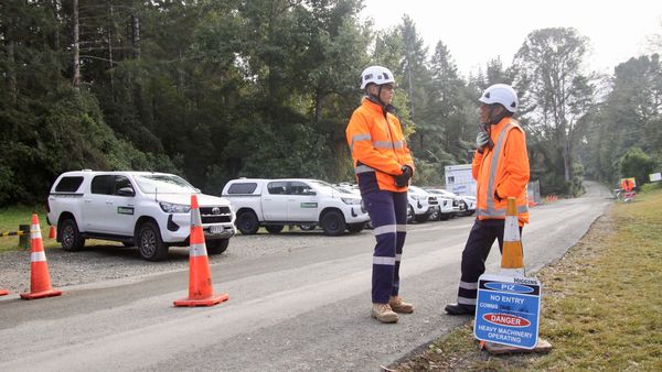 Two people in PPE talking at side of road with utility vehicles parked in the background