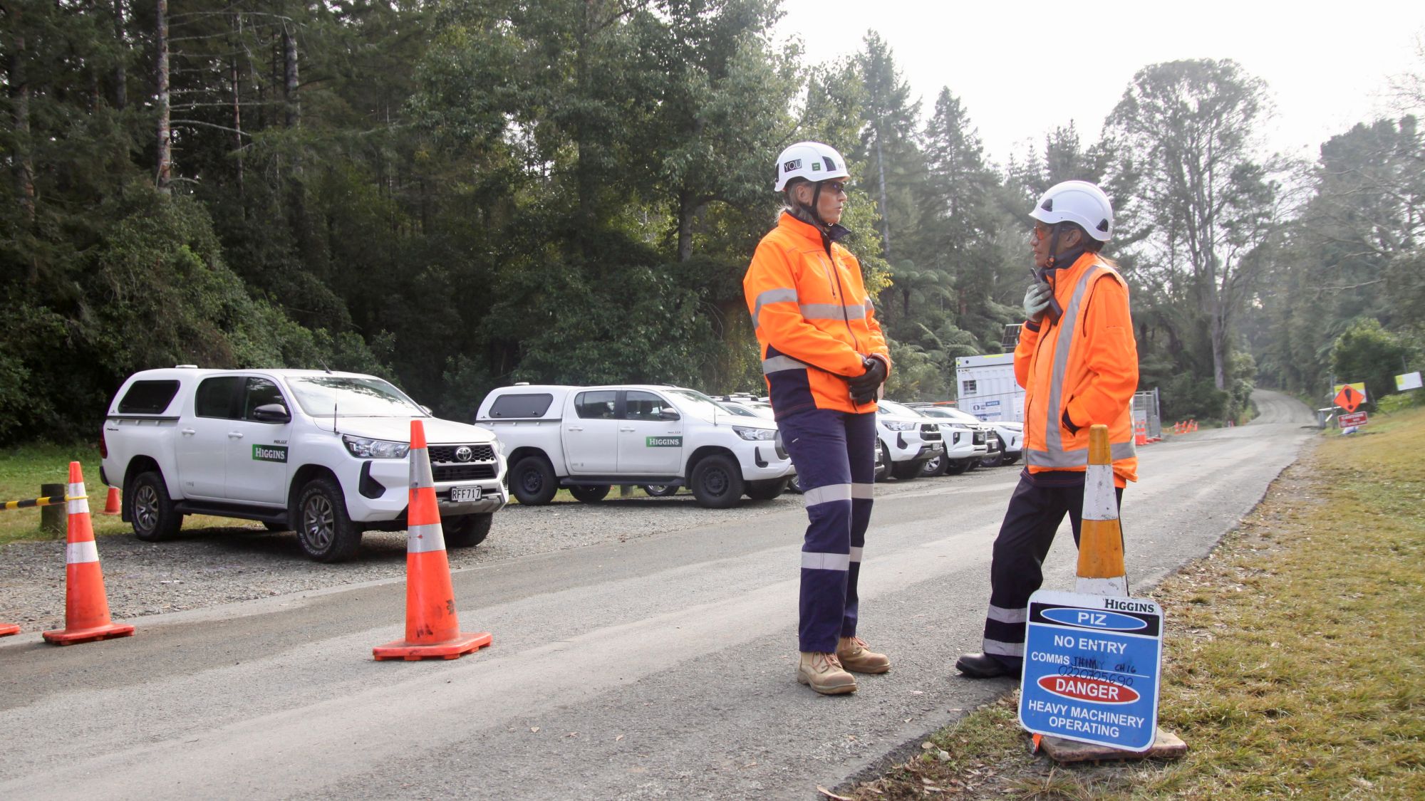 Two people in PPE talking at side of road with utility vehicles parked in the background