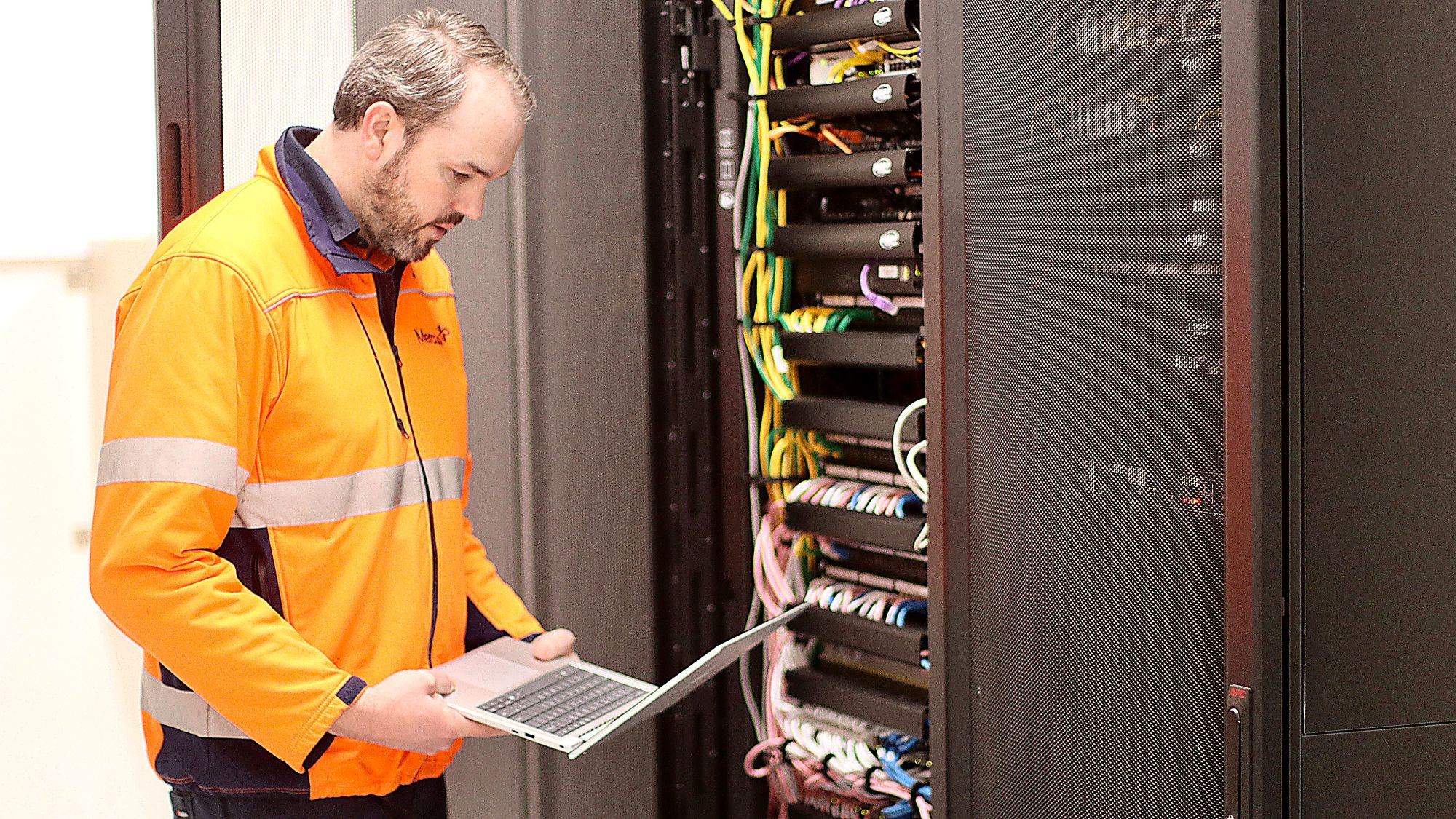 Man in orange hi-viz jacket holding laptop inside a server room with cables in background