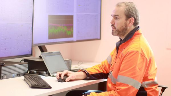 Man in orange hi-viz jacket sitting at a desk operating a mouse with two large screens and a laptop