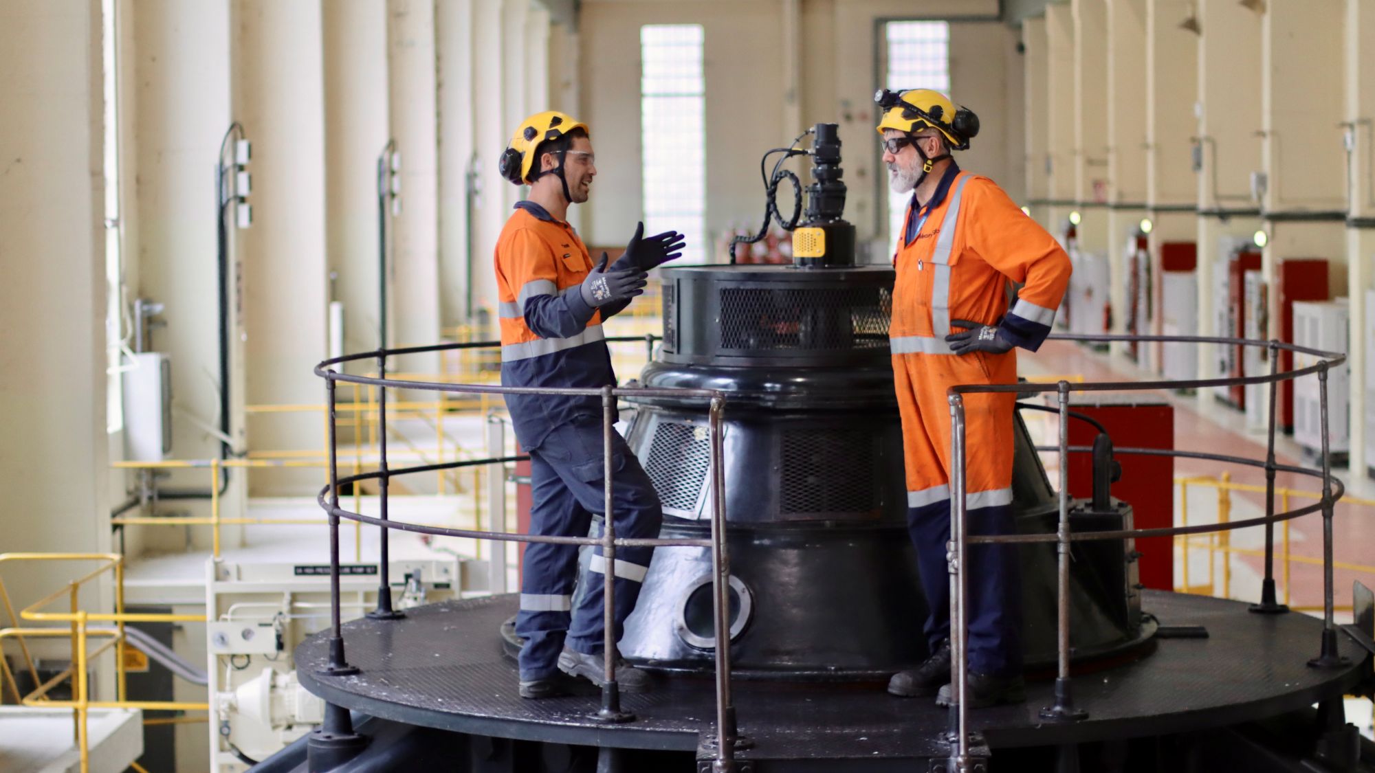 Two men in conversation wearing PPE inside turbine hall at Arapuni Hydro Station
